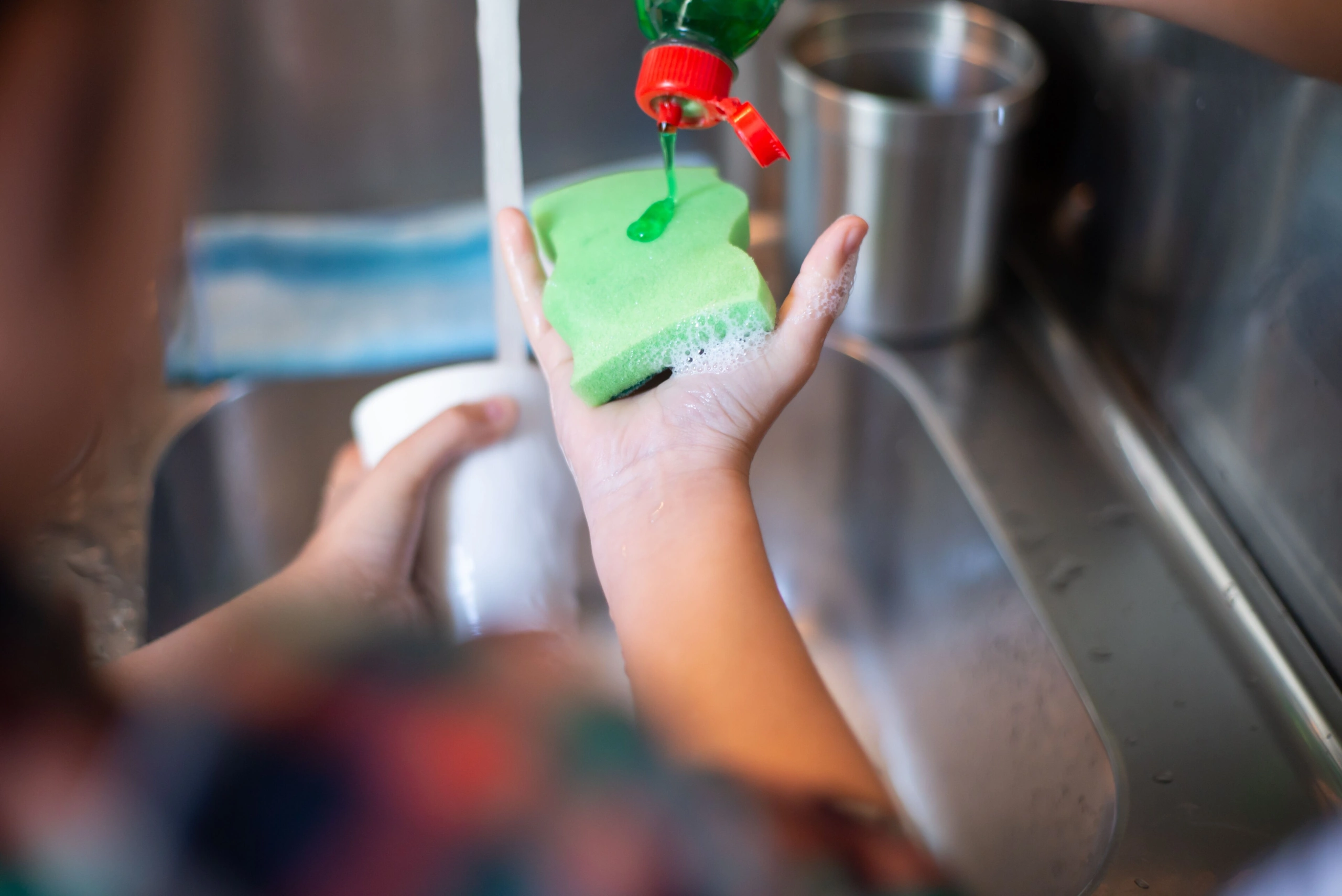 Eine Person gibt Spülmittel auf einen grünen Schwamm, während im Hintergrund Wasser über eine Tasse läuft.