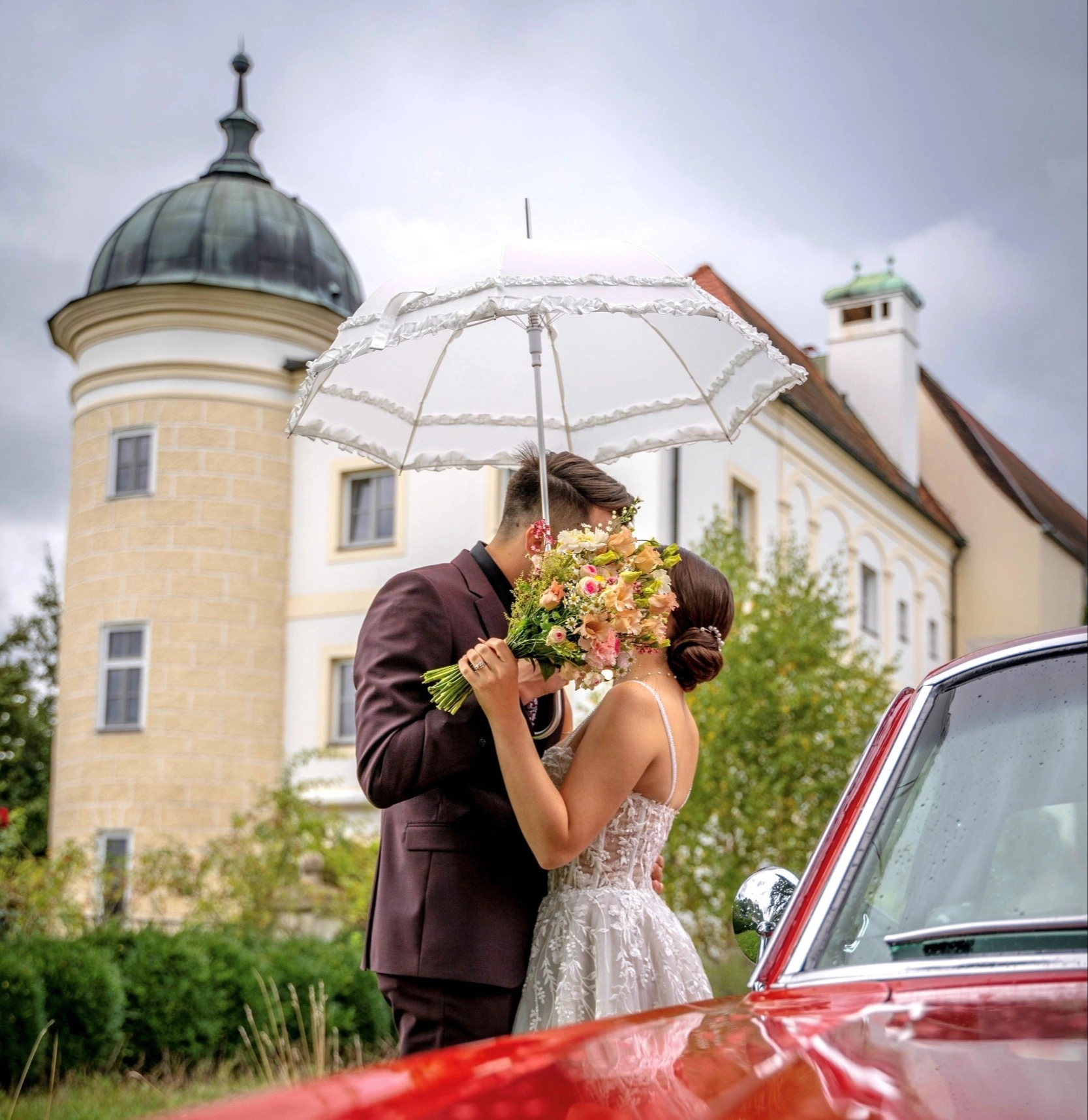 Brautpaar küsst sich unter einem Schirm vor dem Schloss, daneben ein roter Oldtimer